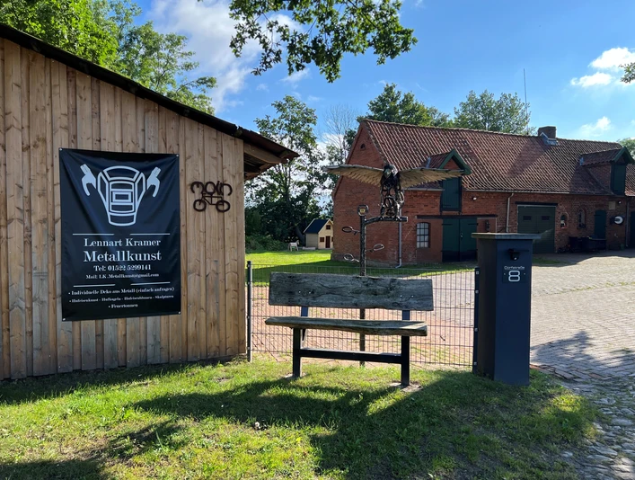 Skulptur Adler Metallene Skulptur eines Adlers auf einem Holzpfosten vor einem Gebäude, umgeben von grünem Gelände.
