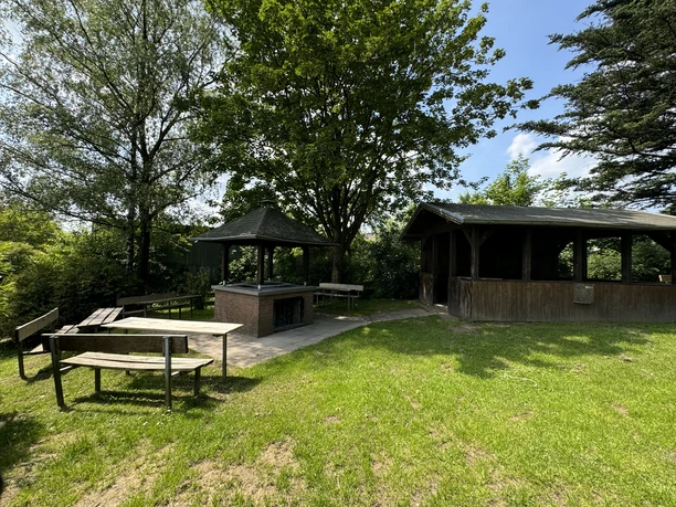 Grillhütte Hohkeppel Grüne Picknick-Wiese mit Holzbänken, Grillbereich und Pavillon, umgeben von Bäumen und strahlendem Himmel.