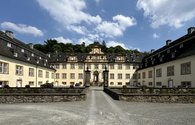 Schloss Ehreshoven Barockes Schloss mit hellgelben Fassaden, symmetrischen Flügeln und zentralem Torbogen, umgeben von Bäumen.
