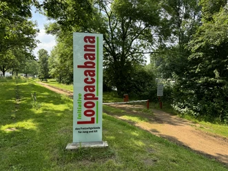 Freizeitanlage Loopacbana Grüner Weg im Park, rechts große Bäume, links Schild mit "Loopacabana" in Rot und Grün.