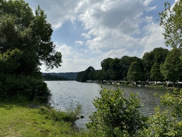 Loopacabana Ein idyllischer See umgeben von üppiger Vegetation und bewaldeten Hügeln unter leicht bewölktem Himmel.