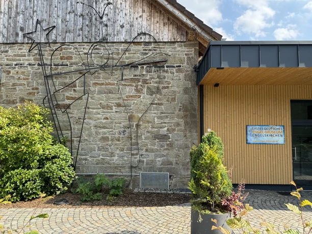 Engel-Museum Eine moderne Wandkunst eines Engels mit Stern aus Metall an einer Steinwand, umgeben von Pflanzen.