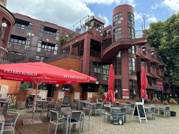 Bürgerhaus Bergischer Löwe Mehrstöckiges Backsteingebäude mit runden Glaselementen, Außengastronomie mit roten Sonnenschirmen.