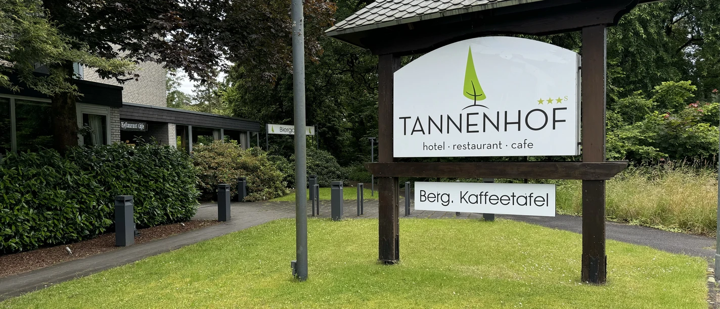 Hotelschild des Tannenhofs auf grüner Wiese, umgeben von Bäumen und einem modernen Gebäude.