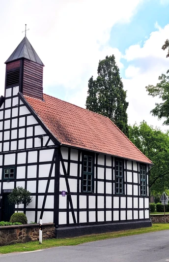 Fachwerkkapelle mit rotem Ziegeldach und kleinem Turm, umgeben von grünen Bäumen an einer Straße.