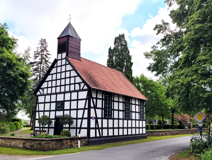 Fachwerkkapelle mit rotem Ziegeldach und kleinem Turm, umgeben von grünen Bäumen an einer Straße.