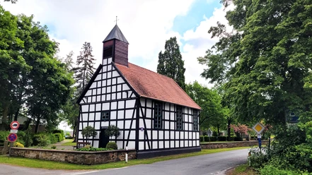 Alte Klus Fachwerkkapelle mit rotem Ziegeldach und kleinem Turm, umgeben von grünen Bäumen an einer Straße.