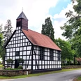 Alte Klus Fachwerkkapelle mit rotem Ziegeldach und kleinem Turm, umgeben von grünen Bäumen an einer Straße.