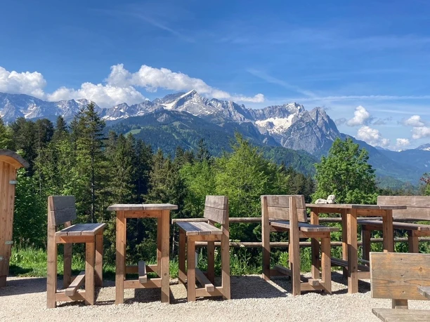 Tannenhütte.jpg Holzstühle und Tische vor einer alpinen Bergkulisse bei sonnigem Wetter.