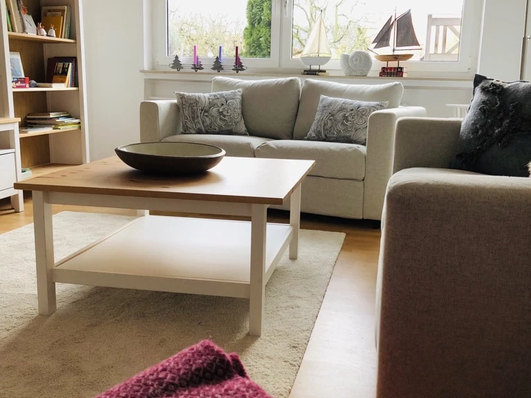 IMG_6222.jpeg Ein helles Wohnzimmer mit zwei Sofas, Couchtisch, Regal, maritimen Dekorationen und großem Fenster.