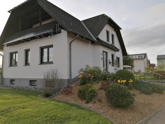 IMG_20240411_190658.jpg Einfamilienhaus mit steilem Dach, hellen Fassaden und gepflegtem Garten mit gelben Blumen.