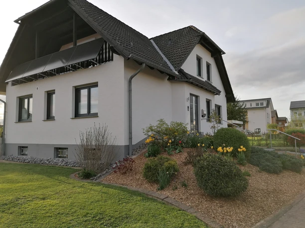 IMG_20240411_190658.jpg Einfamilienhaus mit steilem Dach, hellen Fassaden und gepflegtem Garten mit gelben Blumen.