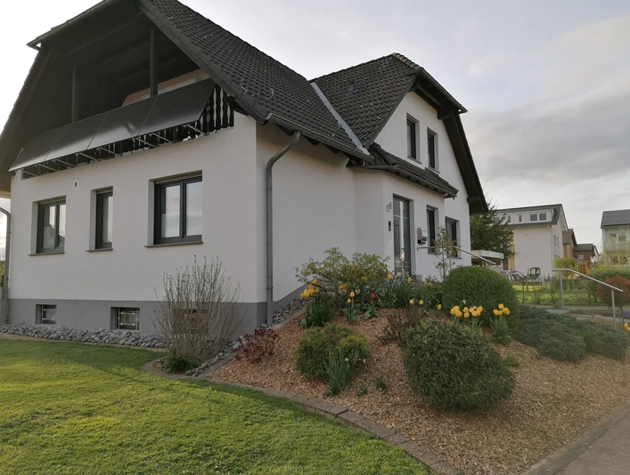 IMG_20240411_190658.jpg Einfamilienhaus mit steilem Dach, hellen Fassaden und gepflegtem Garten mit gelben Blumen.