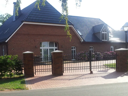 20150806_174627-2.jpg Ein modernes Backsteingebäude mit blauen Dachziegeln und gepflegtem Vorgarten hinter dekorativem Tor.