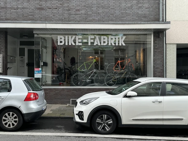 Bike-Fabrik Fahrradgeschäft mit Schaufenster, in dem moderne Fahrräder ausgestellt sind; davor geparkte Autos.