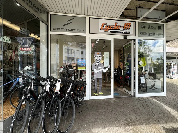 Cycle-M Fahrradgeschäft mit verschiedenen Fahrradmodellen und Zubehör im Schaufenster, Eingang mit Figur.