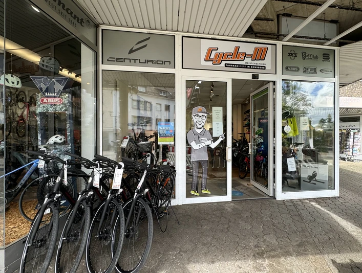 Cycle-M Fahrradgeschäft mit verschiedenen Fahrradmodellen und Zubehör im Schaufenster, Eingang mit Figur.