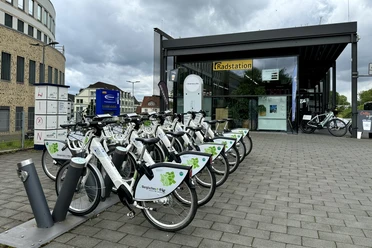 E-Bike Verleihstation Bergisch Gladbach Leihfahrräder vor einer modernen Radstation auf gepflastertem Platz, umgeben von Büros und Wolken.
