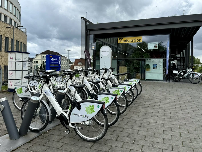E-Bike Verleihstation Bergisch Gladbach Leihfahrräder vor einer modernen Radstation auf gepflastertem Platz, umgeben von Büros und Wolken.