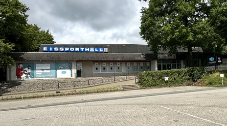Eissporthalle Bergisch Gladbach Eine graue Eissporthalle mit blauem Schild und umliegender Natur unter bewölktem Himmel.