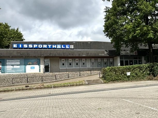 Eissporthalle Bergisch Gladbach Eine graue Eissporthalle mit blauem Schild und umliegender Natur unter bewölktem Himmel.