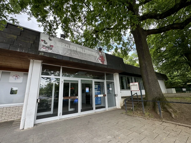 Eissportarena Eingangsbereich eines Gebäudes mit großen Glasfenstern, Willkommensschild und einem großen Baum daneben.