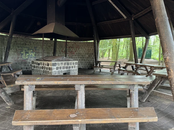 Grillhütte Holzbänke und Tische unter einem runden hölzernen Pavillon mit Grillplatz in einem Waldgebiet.