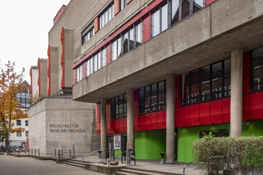 University of Music and Dance Striking building of the Cologne University of Music and Dance with red and green accents on the façade, surrounded by trees and urban architecture.