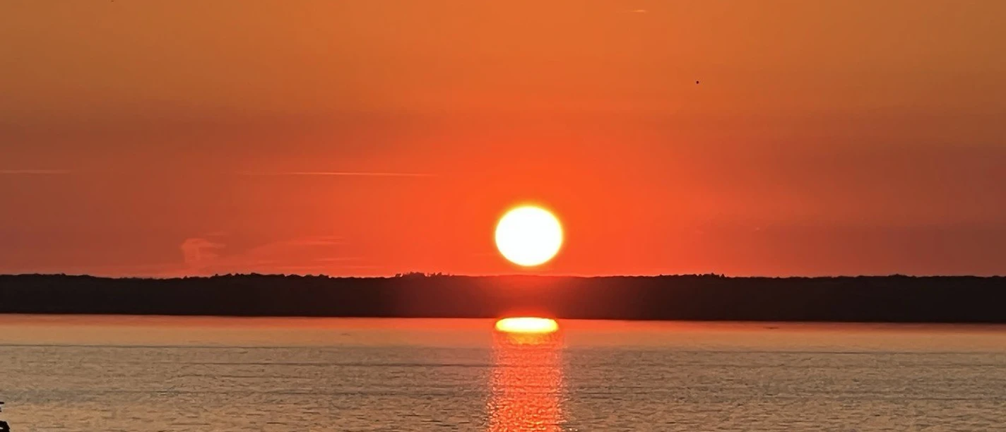 Bild 2.jpg Leuchtender Sonnenuntergang über ruhigem Wasser, reflektiert kräftige Orangetöne am klaren Himmel.