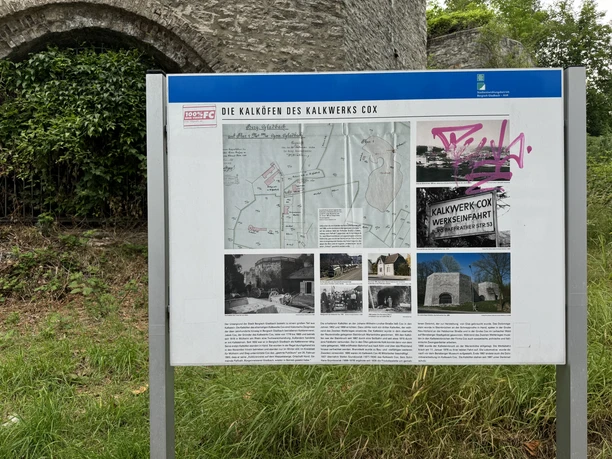 Die Kalköfen des Kalkwerks Cox Informationstafel mit Texten und Bildern über das historische Kalkwerk Cox, umgeben von grünem Laub.