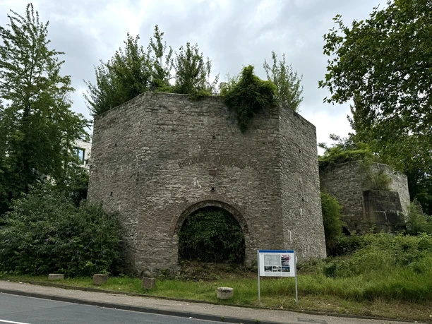 Kalköfen Alte, teils bewachsene Befestigungsruine aus Stein mit runder Form, umgeben von Bäumen und Schildern.