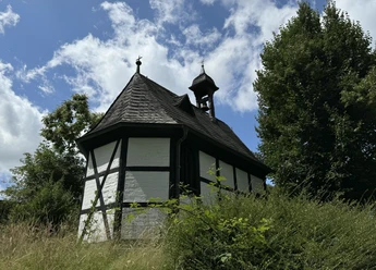 Barbarakapelle Fachwerkkapelle in ländlicher Umgebung, umgeben von grüner Vegetation und Bäumen unter blauem Himmel.