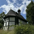 Barbarakapelle Fachwerkkapelle in ländlicher Umgebung, umgeben von grüner Vegetation und Bäumen unter blauem Himmel.