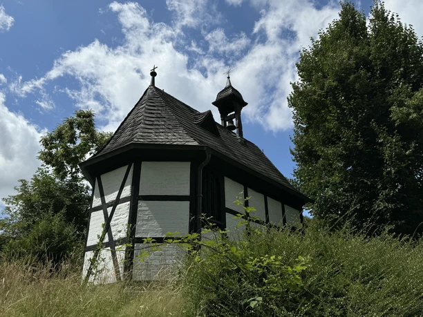 Barbarakapelle Fachwerkkapelle in ländlicher Umgebung, umgeben von grüner Vegetation und Bäumen unter blauem Himmel.