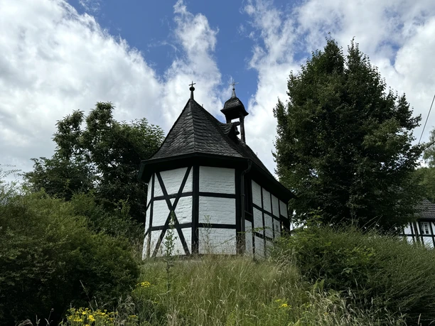 Barbarakapelle Kleine Fachwerkkapelle mit Schieferdach, umgeben von Bäumen und Wiesen unter bewölktem Himmel.