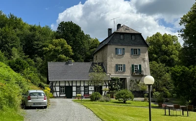 Burghaus Hellenthal Historische Villa mit Fachwerk-Nebengebäude in grüner, ländlicher Umgebung unter teils bewölktem Himmel.