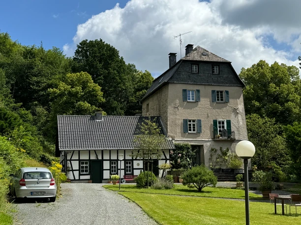 Burghaus Hellenthal Historische Villa mit Fachwerk-Nebengebäude in grüner, ländlicher Umgebung unter teils bewölktem Himmel.