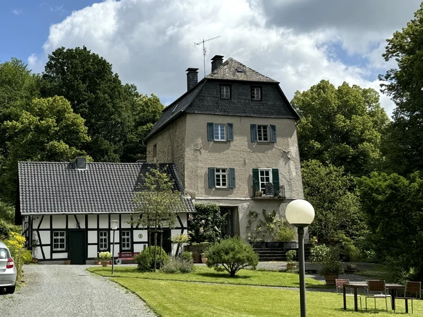 Burghaus Hellenthal Ein historisches, dreistöckiges Steinhaus mit Fachwerkgebäude, umgeben von Bäumen und gepflegtem Garten.