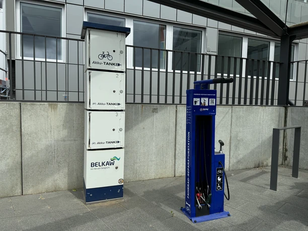 Ladesäule Fahrrad-Ladestation und Reparaturstation unter einem Vordach mit Betonwand und modernem Gebäude im Hintergrund.