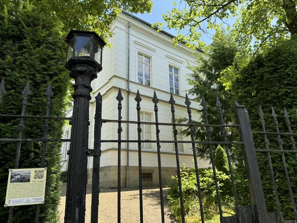 Villa Longrée - Straßenansicht Historische weiße Villa mit schmiedeeisernem Zaun, von grünen Bäumen und Sträuchern umgeben.