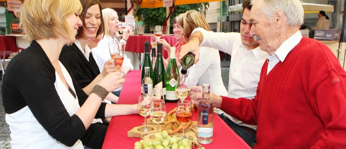 Eine Gruppe von Personen sitzt an einem rot gedeckten Tisch im Freien, trinkt Wein und lacht zusammen.