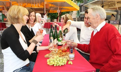 WeinSommer Aachen Eine Gruppe von Personen sitzt an einem rot gedeckten Tisch im Freien, trinkt Wein und lacht zusammen.