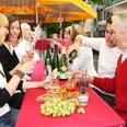 WeinSommer Aachen Eine Gruppe von Personen sitzt an einem rot gedeckten Tisch im Freien, trinkt Wein und lacht zusammen.