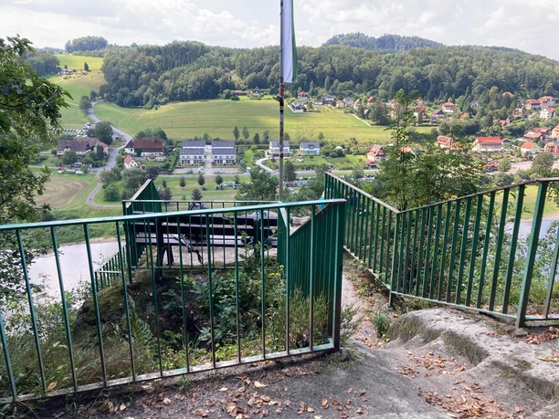 "Kleine_Bastei"_uitzichtspunt Groene balustrades leiden naar een uitkijkplatform met een bankje, op de achtergrond een landelijk landschap met huizen en heuvels onder een bewolkte hemel.