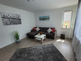 Heller Wohnraum mit grauem Sofa, Couchtisch, Wandbildern, einem Teppich und hellem Holzfußboden.