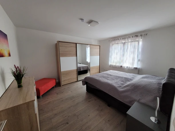 Moderner Schlafraum mit Doppelbett, großem Schrank, Nachttisch und rotem Sitzhocker, hell dekoriert.