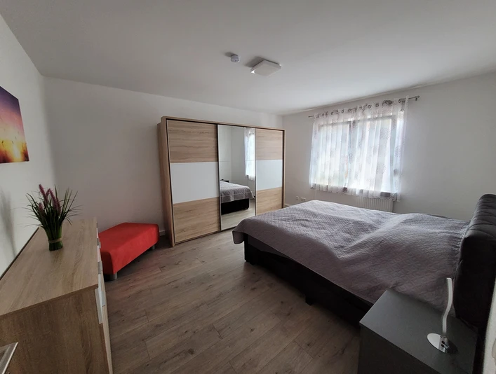 Moderner Schlafraum mit Doppelbett, großem Schrank, Nachttisch und rotem Sitzhocker, hell dekoriert.