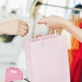 Eine rosa Einkaufstasche wird zwischen zwei Personen vor einer bunten, mit Kleidung gefüllten Kulisse übergeben.