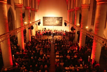In einem beleuchteten Kirchenraum steht ein Chor vor vollen Bänken unter gotischen Bögen.