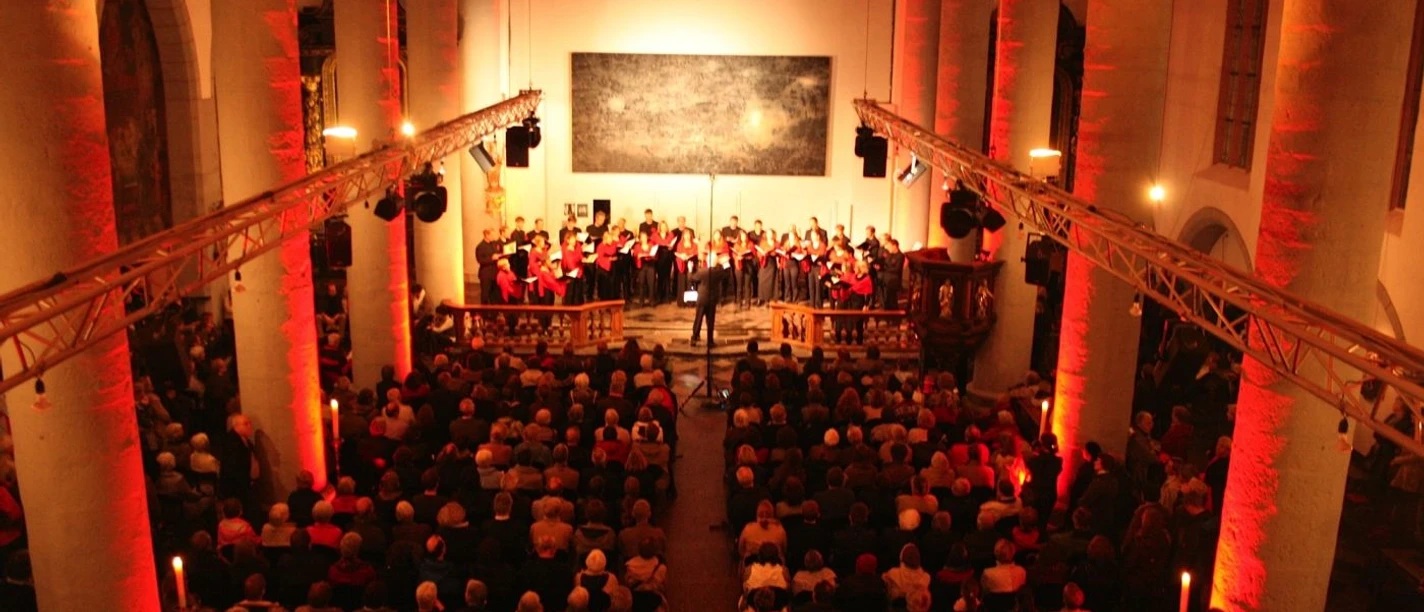 In einem beleuchteten Kirchenraum steht ein Chor vor vollen Bänken unter gotischen Bögen.